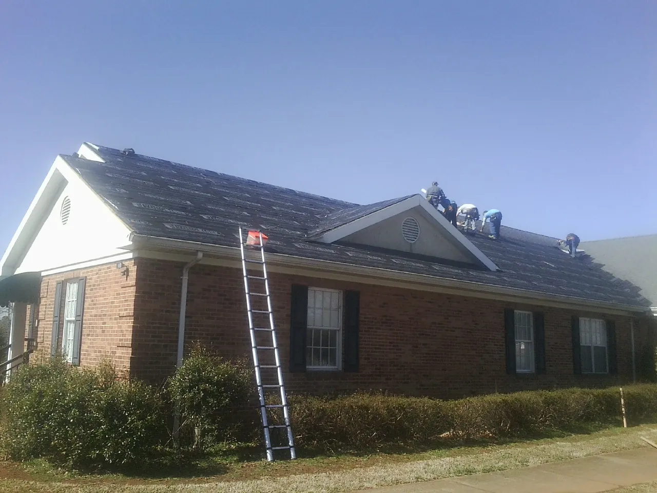 Crew tearing shingles off a brick ranch roof with a ladder set — Emergency Roof Repair work by our crew in Moravia