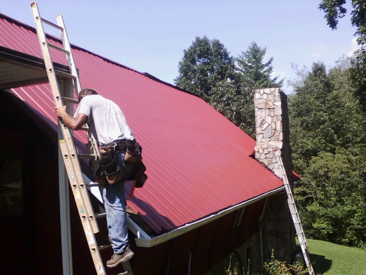 Roofer on a ladder beside a red metal roof and stone chimney — Soffit & Fascia Repair by Roofing Brothers in Moravia
